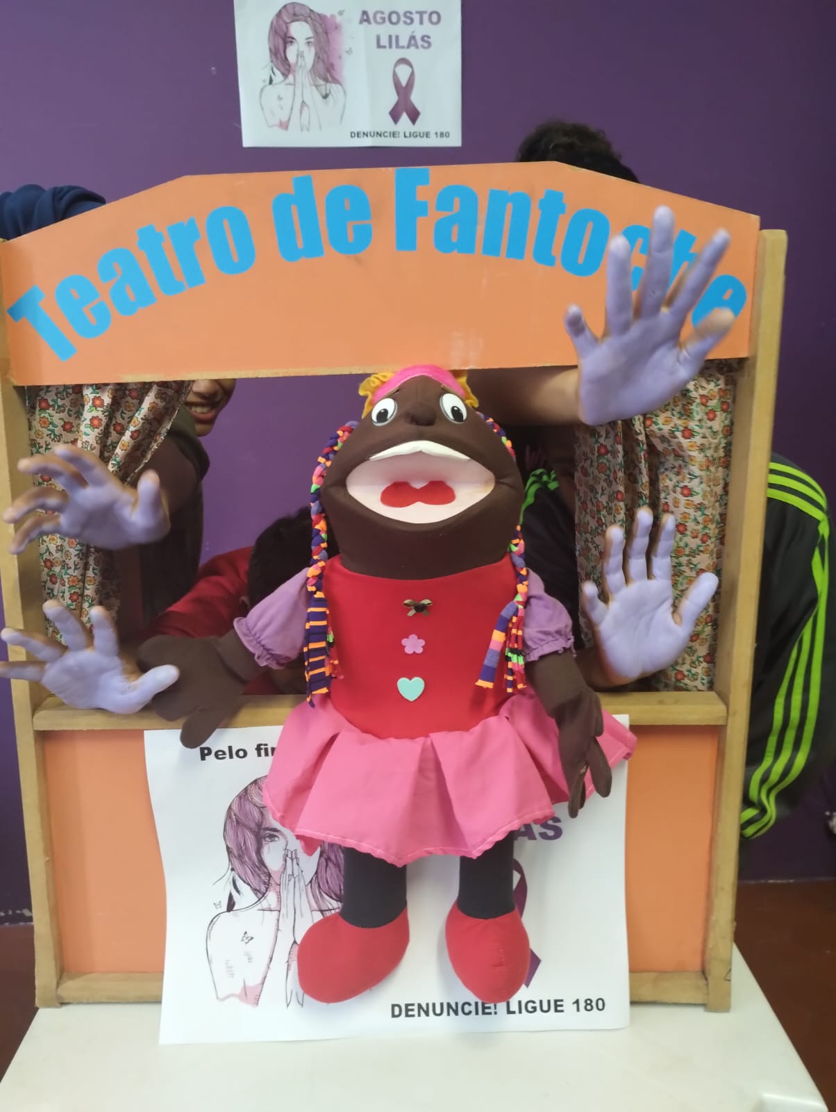 Na imagem, um teatro de fantoches montado, com um boneco na frente, e ao lado, diversas mãos de crianças pintadas de roxo